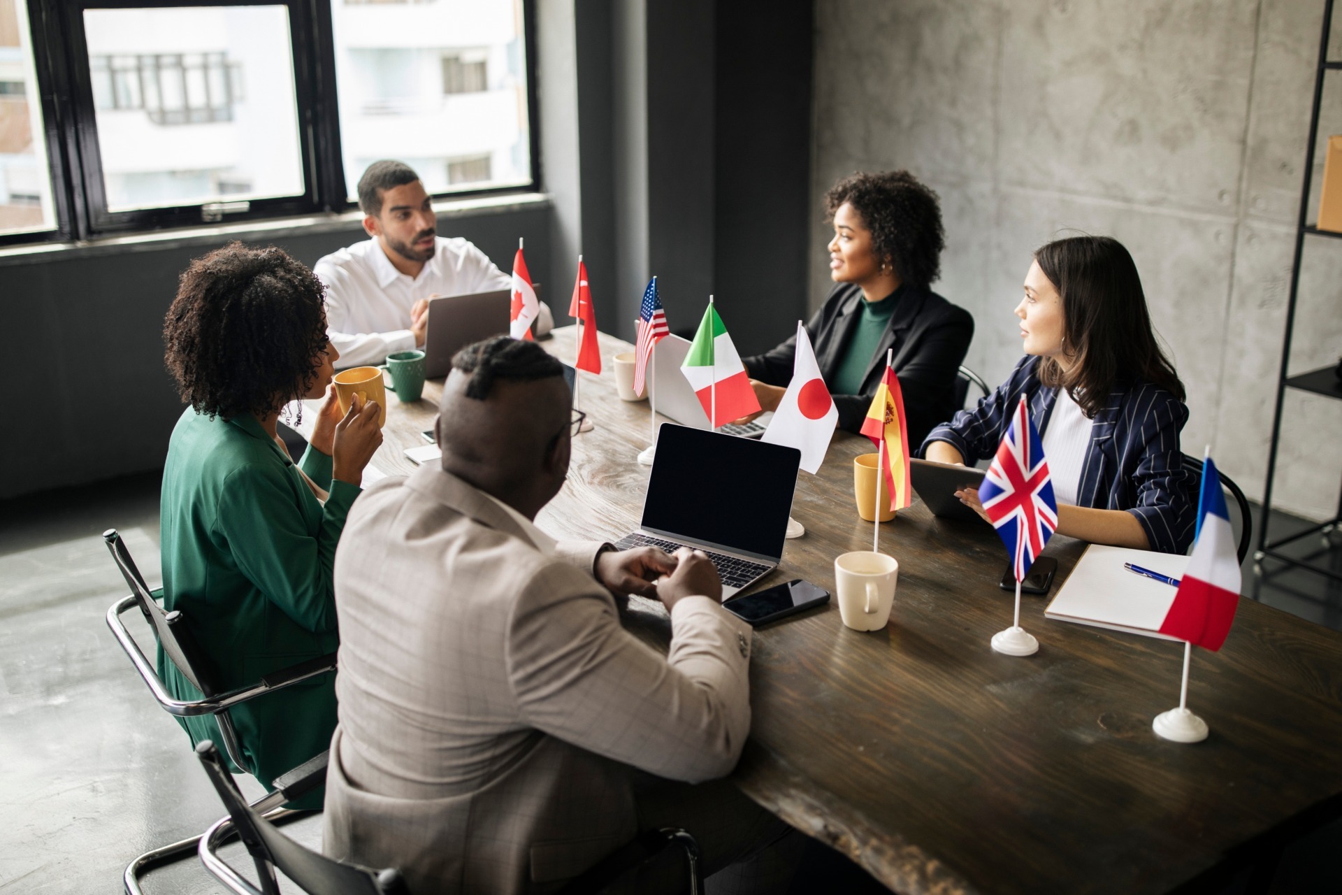 A multicultural team in a meeting around a wooden table lined with small national flags.
