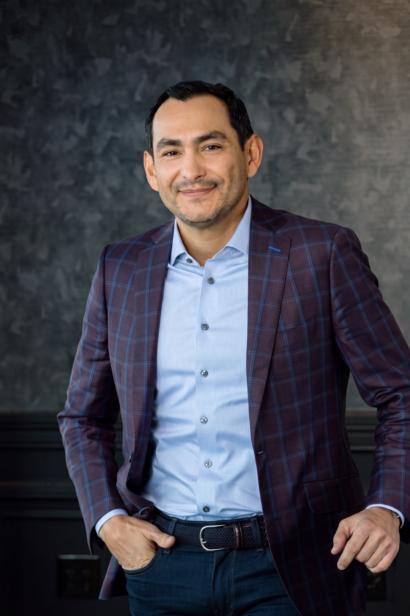 Andres Ceballos, in a dark plaid blazer over a light blue shirt, one hand in his pocket, against a mottled grey studio backdrop.