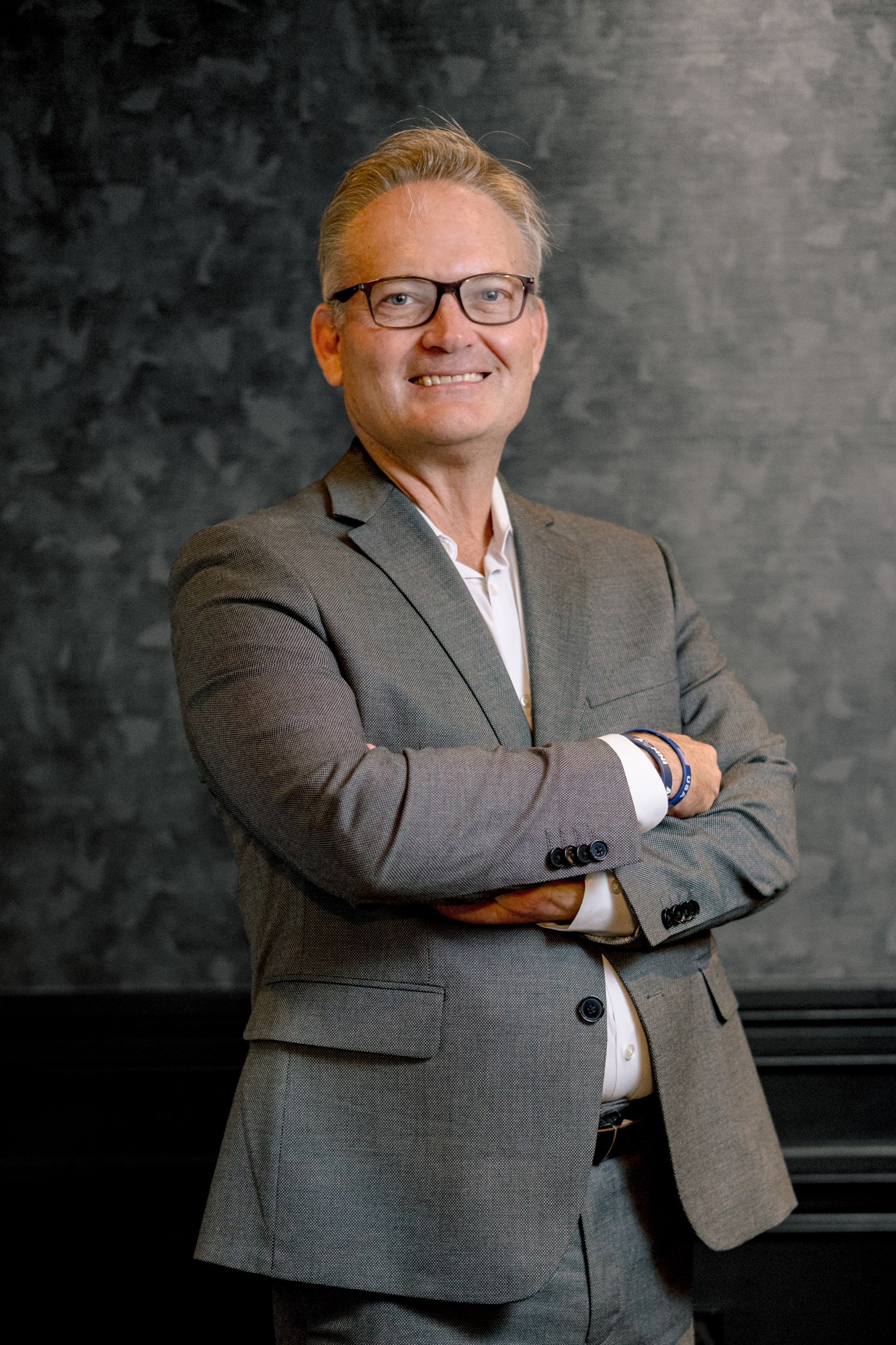 David Lindsey, in a grey suit and glasses, arms crossed, against a mottled grey studio backdrop.