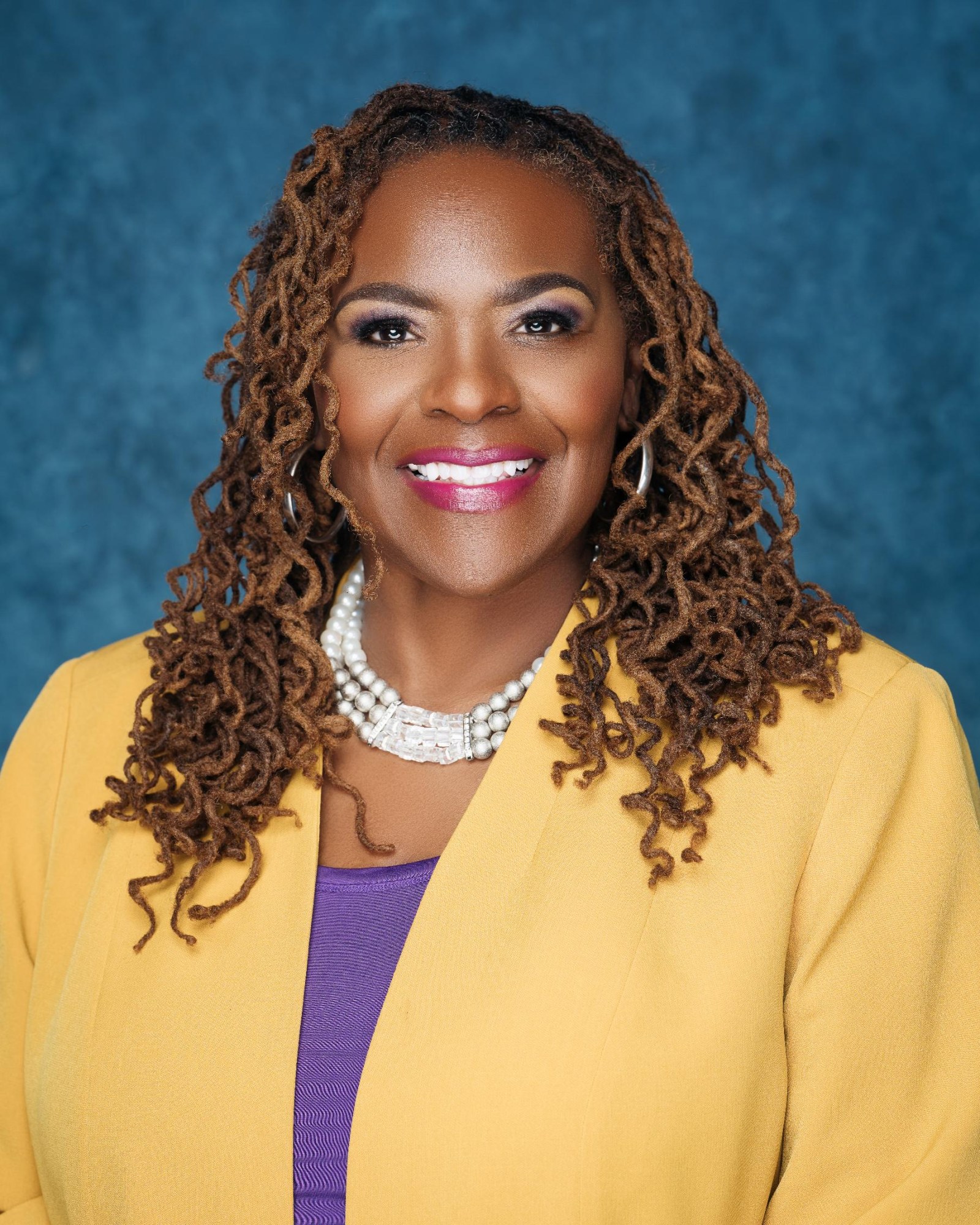 Deloria Nelson, in a yellow jacket over a purple top with a pearl necklace, long copper locs, against a blue studio backdrop.