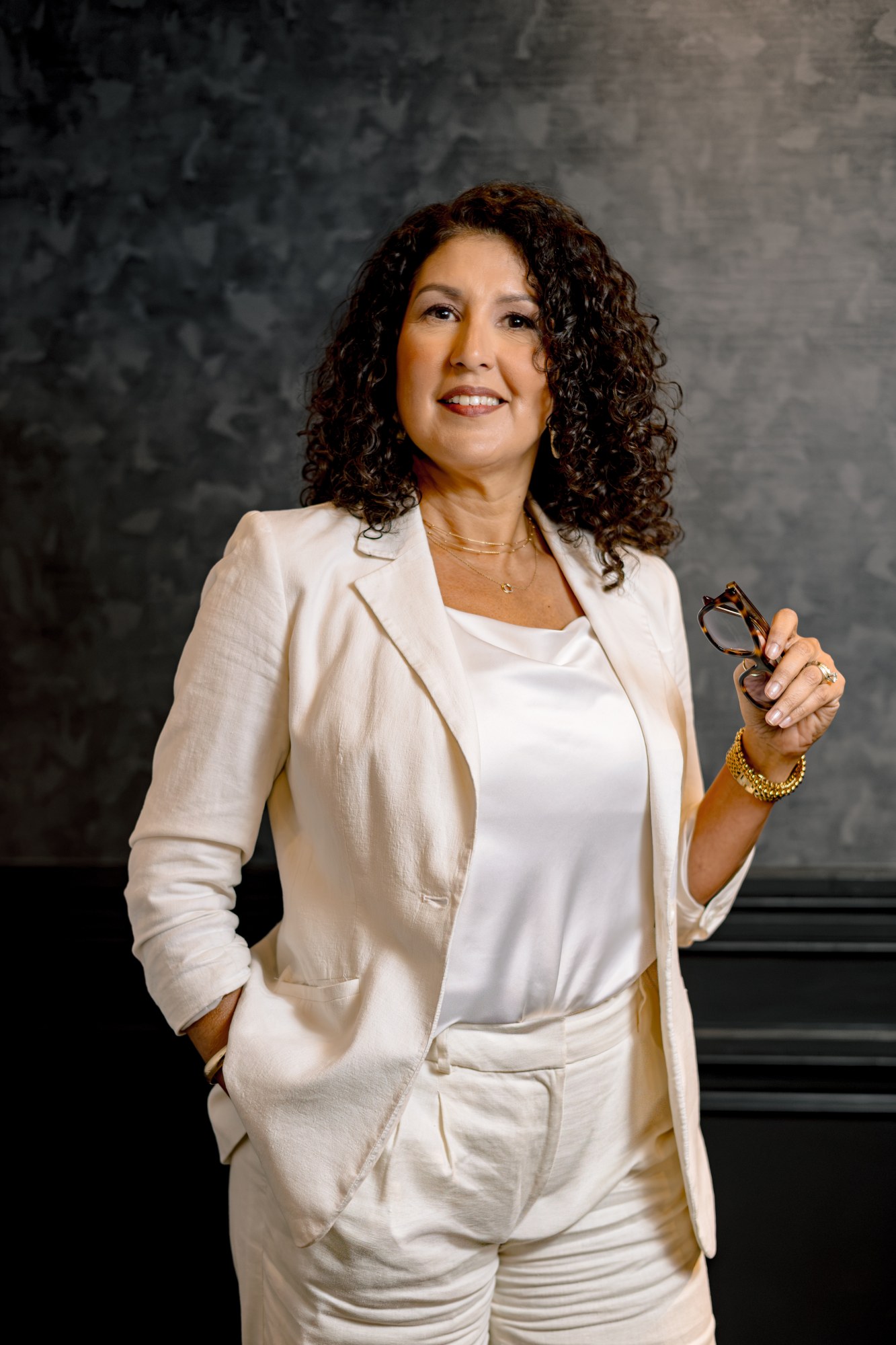 Denise Musselwhite, in a white suit over a white top, dark curls, sunglasses in hand, against a mottled grey studio backdrop.