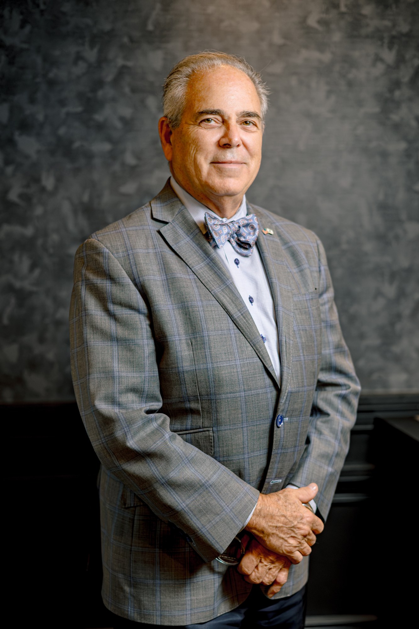 James Gelfand, in a grey check jacket and patterned bow tie, hands clasped, against a mottled grey studio backdrop.