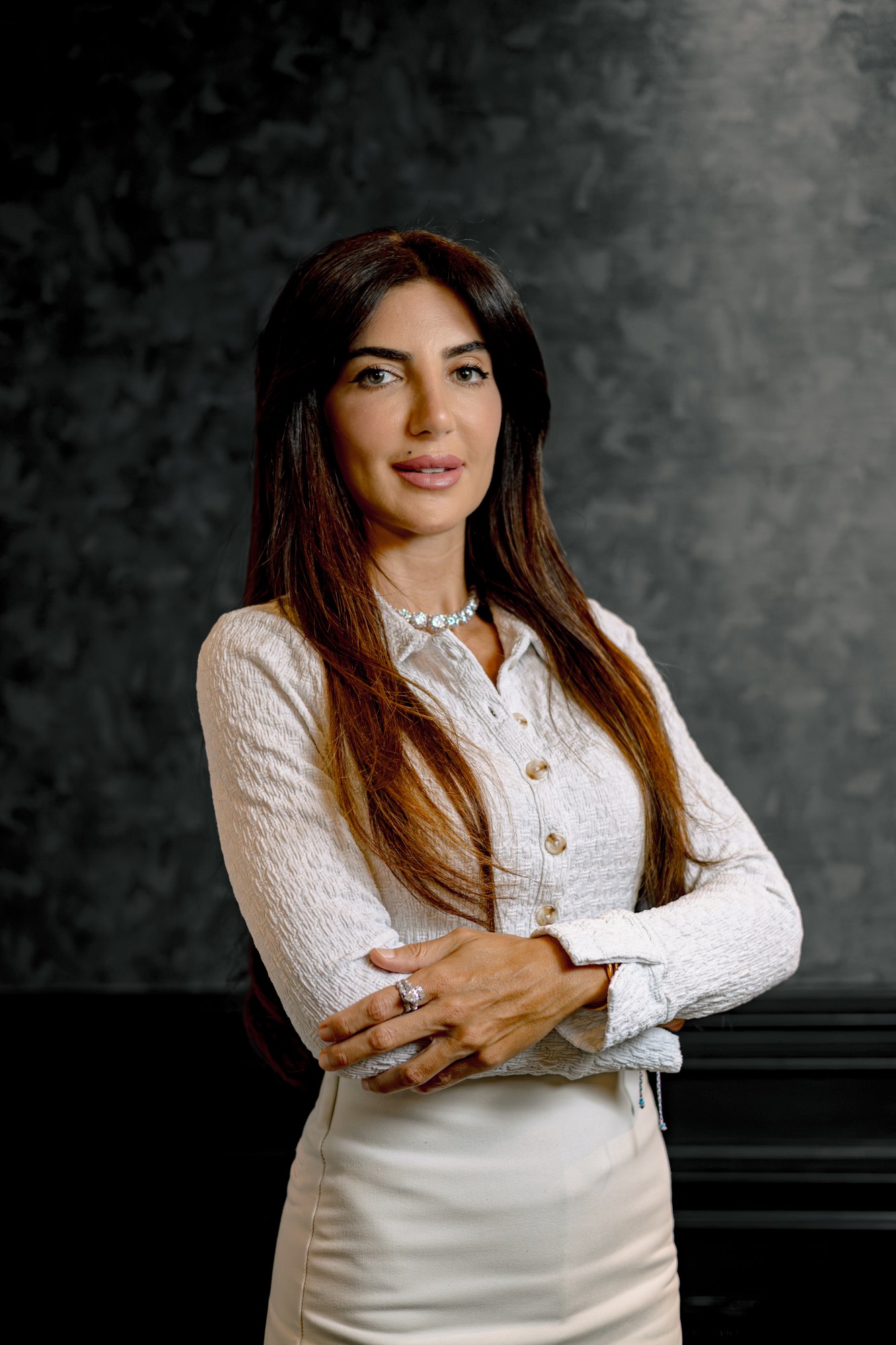 Maria Helena Maroun, in a white textured blouse and white skirt, long dark hair, arms crossed, against a mottled grey studio backdrop.