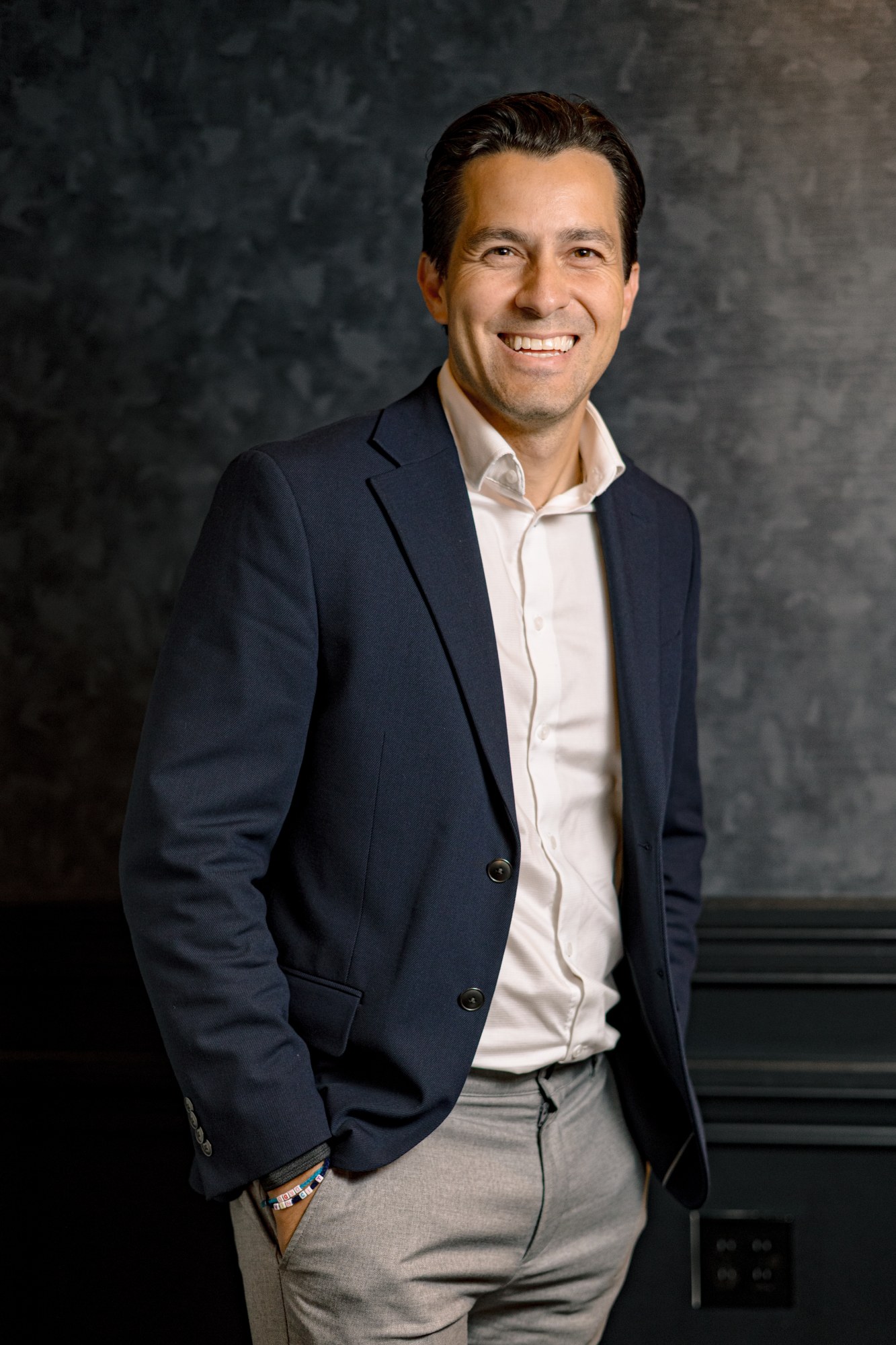 Ricardo Matte, in a navy blazer over an open white shirt, one hand in his pocket, against a mottled grey studio backdrop.