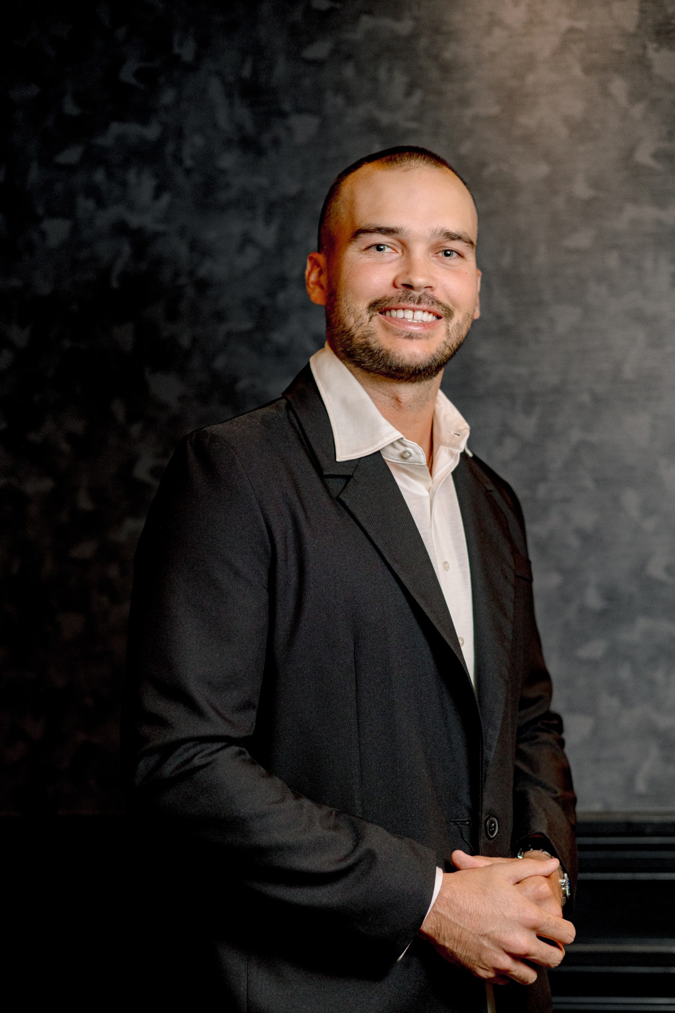 Taylor Akers, in a black jacket over an open white shirt, against a mottled grey studio backdrop.