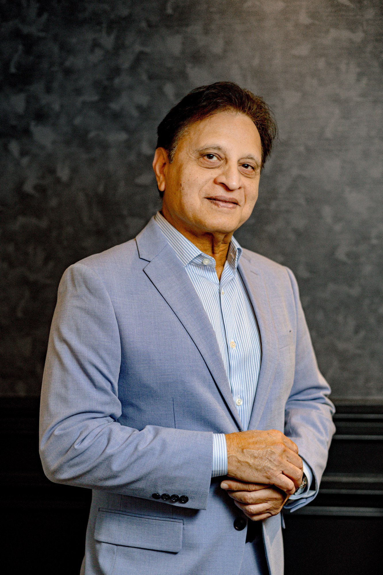 Vincent Desai, in a light blue suit over a pale blue shirt, hands clasped, against a mottled grey studio backdrop.