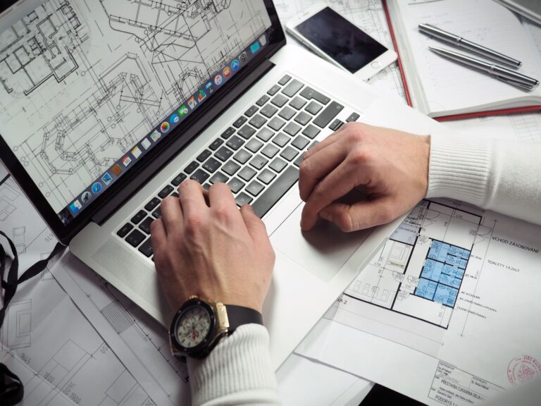 Hands typing on a laptop over a desk spread with architectural drawings