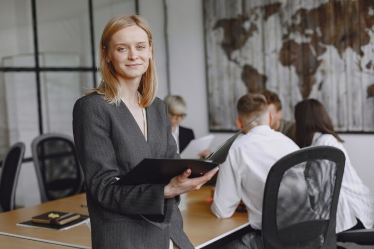 A businesswoman holding a folder, standing before colleagues at a meeting table