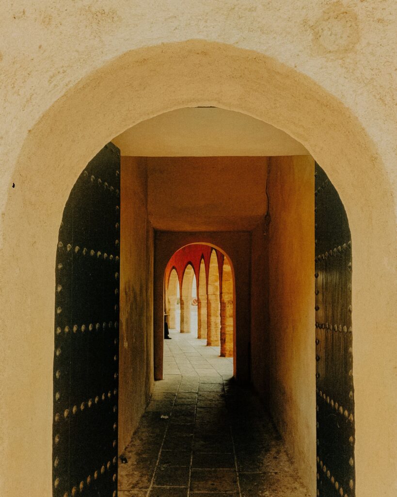 A pale arched gateway opens onto a passage between studded dark doors, leading to a row of sunlit red arches.