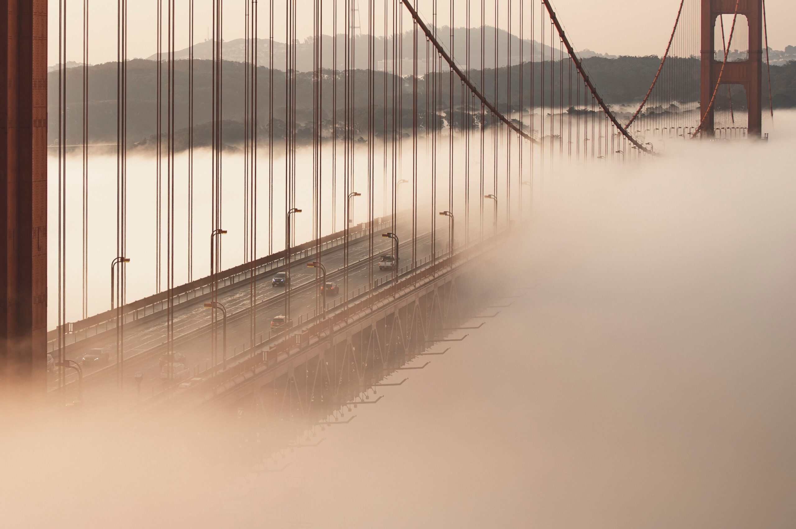 A red suspension bridge disappears into low fog, cars on its deck and hills rising beyond the cables.