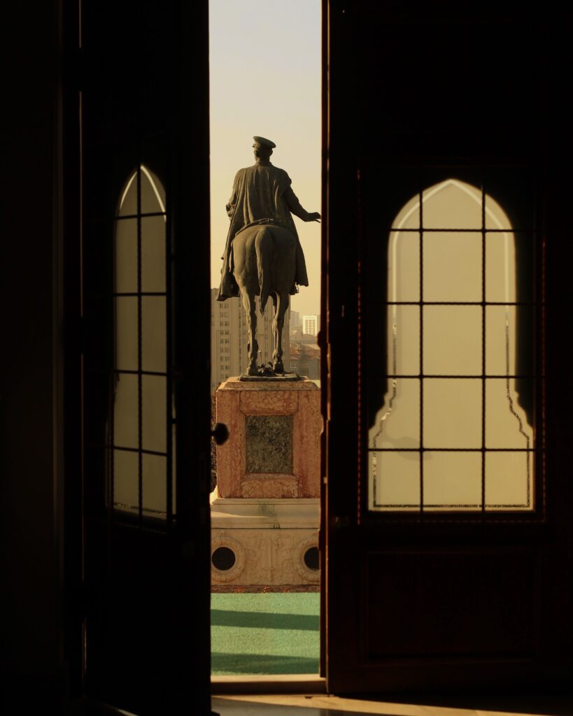 Through a pair of open wooden doors with arched windows, an equestrian statue on a stone plinth stands against a hazy city sky.