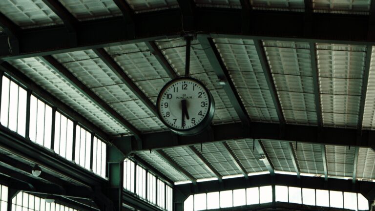 A large station clock hangs from the steel trusses of a skylit hall roof.