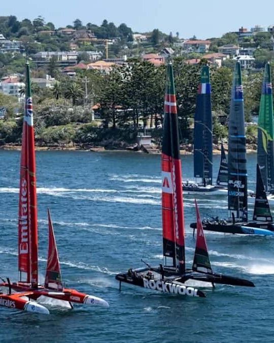 SailGP's fleet racing close to a wooded shoreline lined with houses, Great Britain's red boat at the front