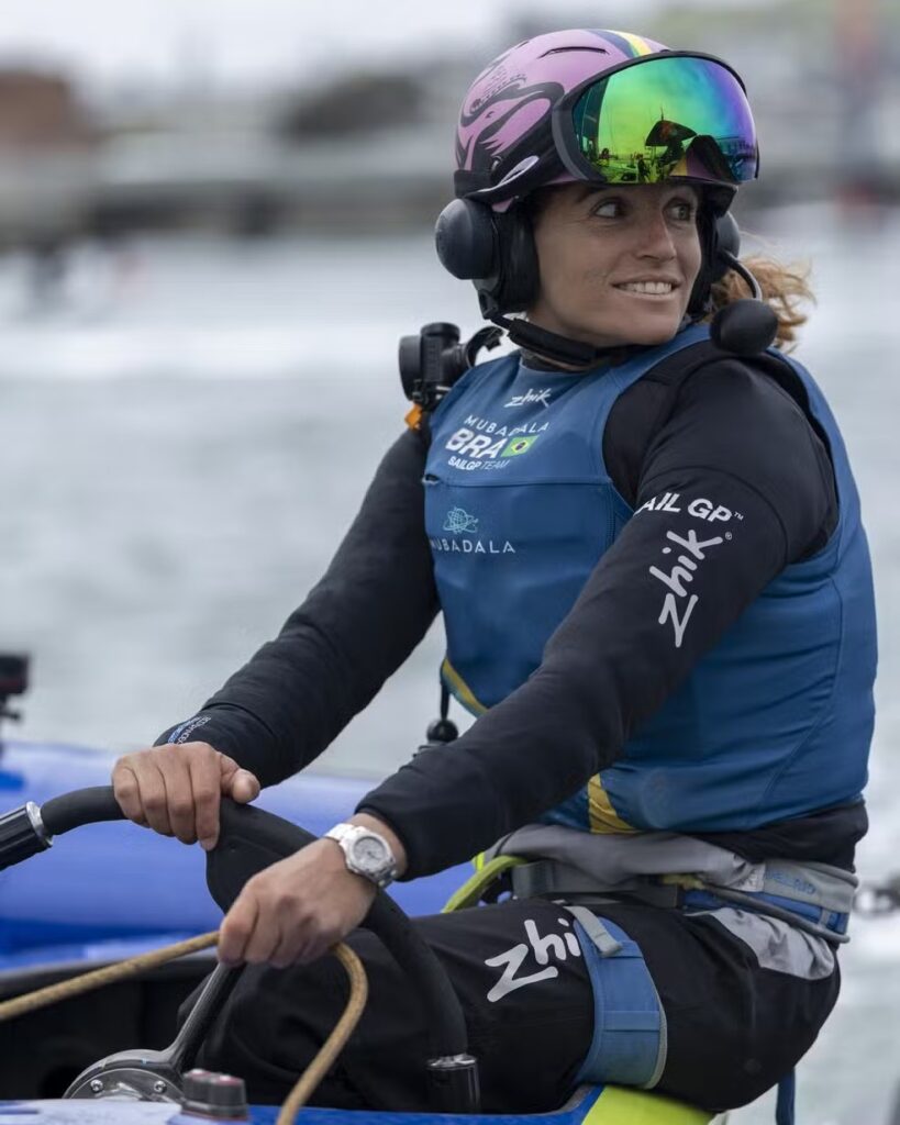A sailor in a Mubadala Brazil SailGP Team vest and mirrored goggles at the wheel of an F50