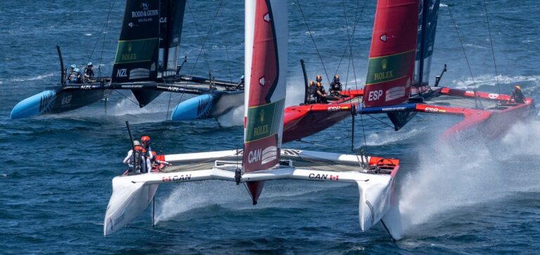 Three SailGP F50 catamarans foiling at speed, Canada's white boat ahead of New Zealand and Spain, spray streaming off the hulls