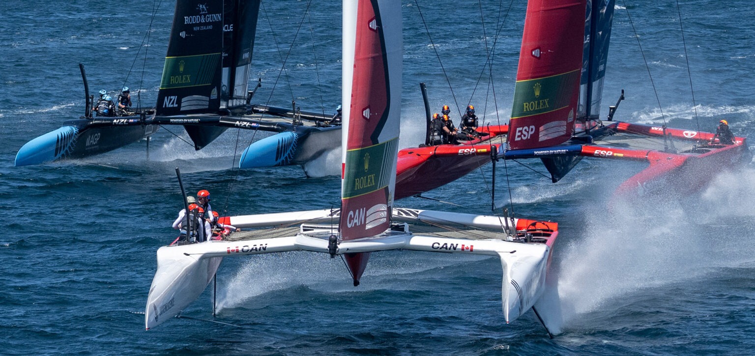 Three SailGP F50 catamarans foiling at speed, Canada's white boat ahead of New Zealand and Spain, spray streaming off the hulls