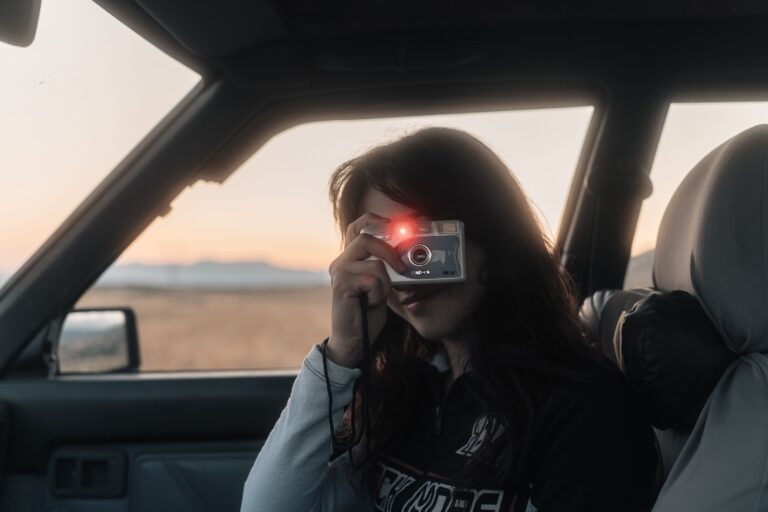A young woman in a car at dusk holds a point-and-shoot film camera to her eye, a dry plain and low hills glowing in the window behind her.