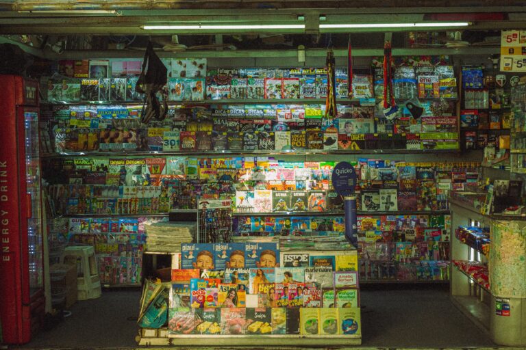 A newsstand lit by fluorescent tubes, its shelves packed floor to ceiling with magazines and comics.